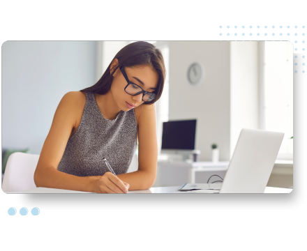 A focused woman with glasses writes in a notebook at a desk, with a laptop open. Symbolizing digital transformation and training in HR technology.