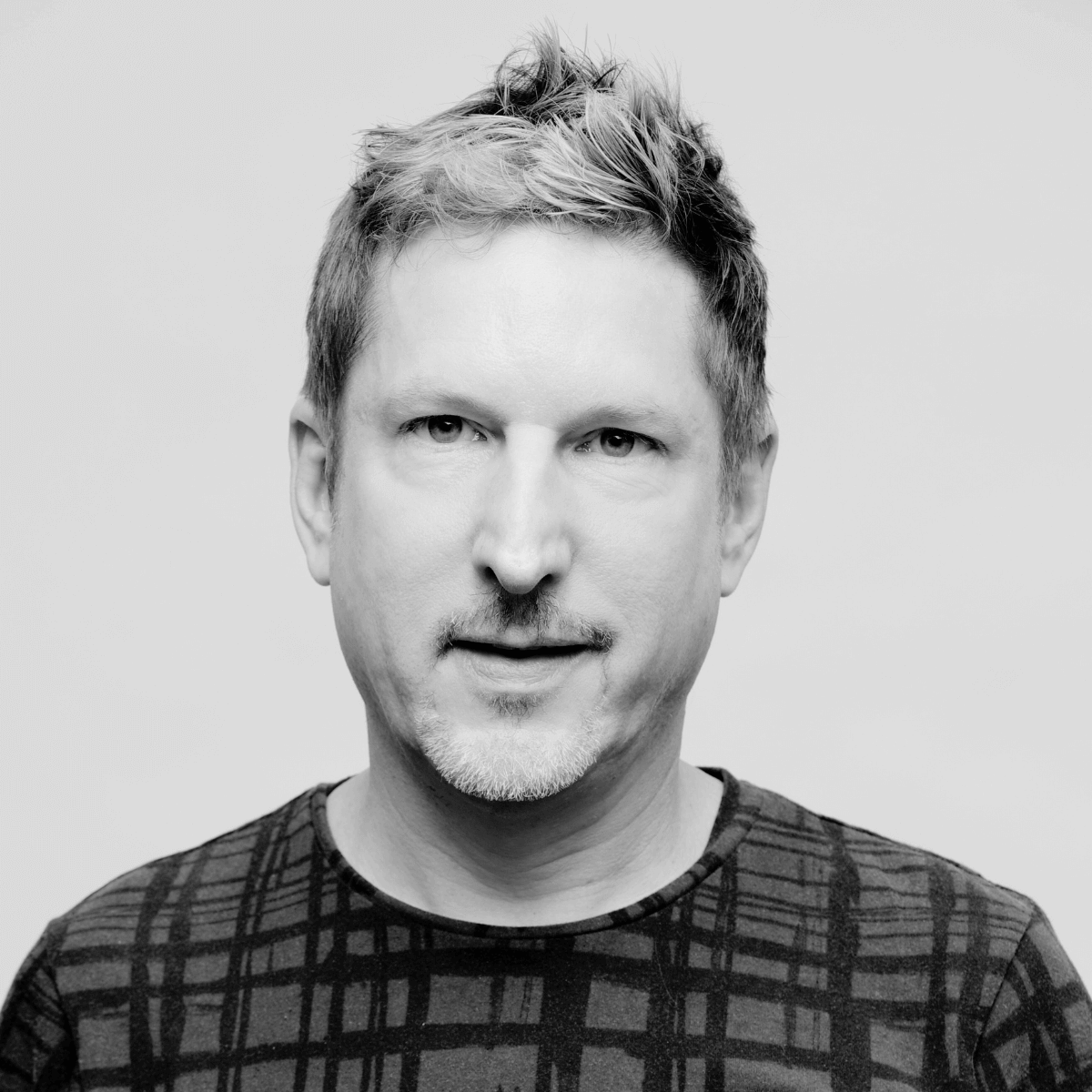Black and white portrait of a man with short, styled hair and a subtle beard, wearing a patterned shirt. He gazes directly, conveying a confident tone.