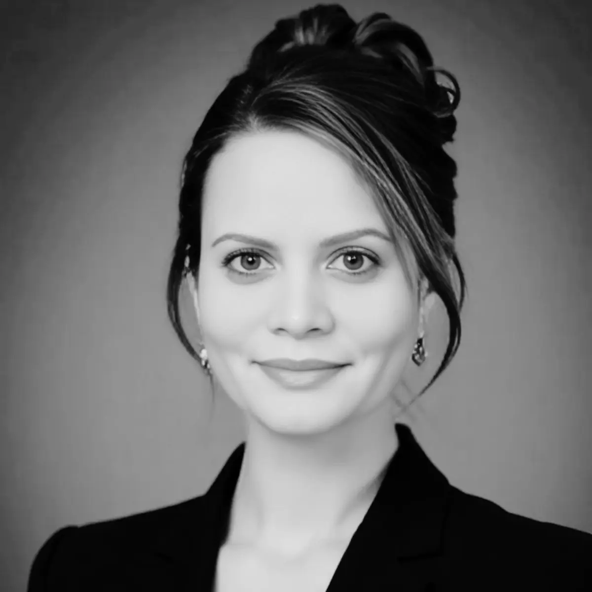 Black and white portrait of a woman with wavy hair smiling warmly. She wears a blazer with a decorative brooch. Neutral background adds elegance.