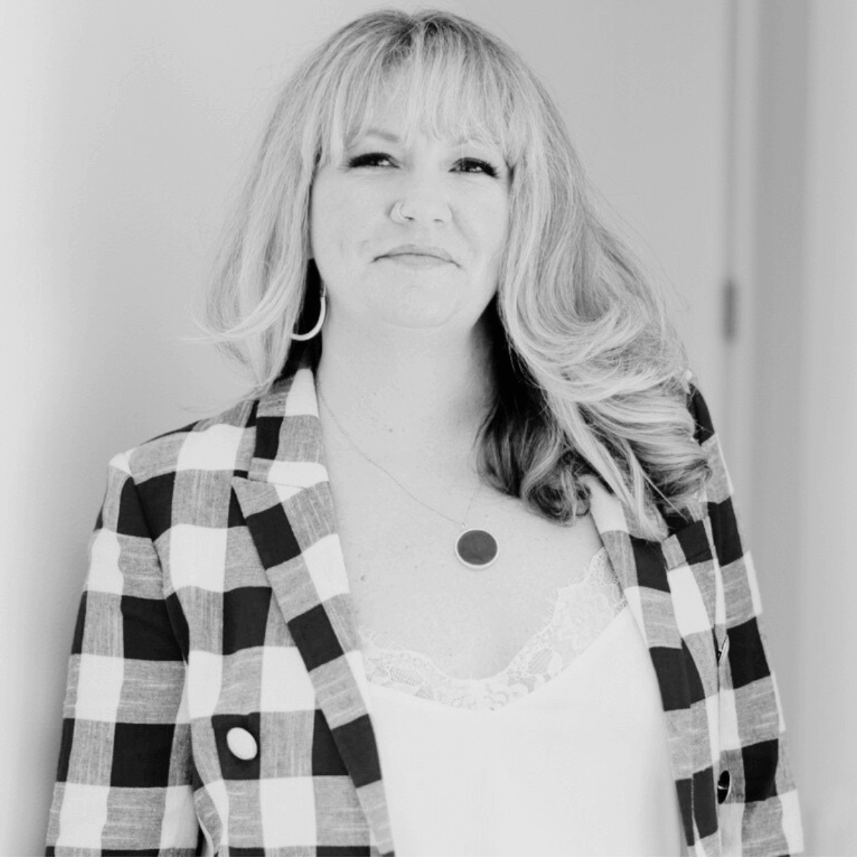 Woman with long, wavy hair smiles confidently, wearing a plaid blazer and necklace. The black and white photo exudes a professional yet relaxed tone.