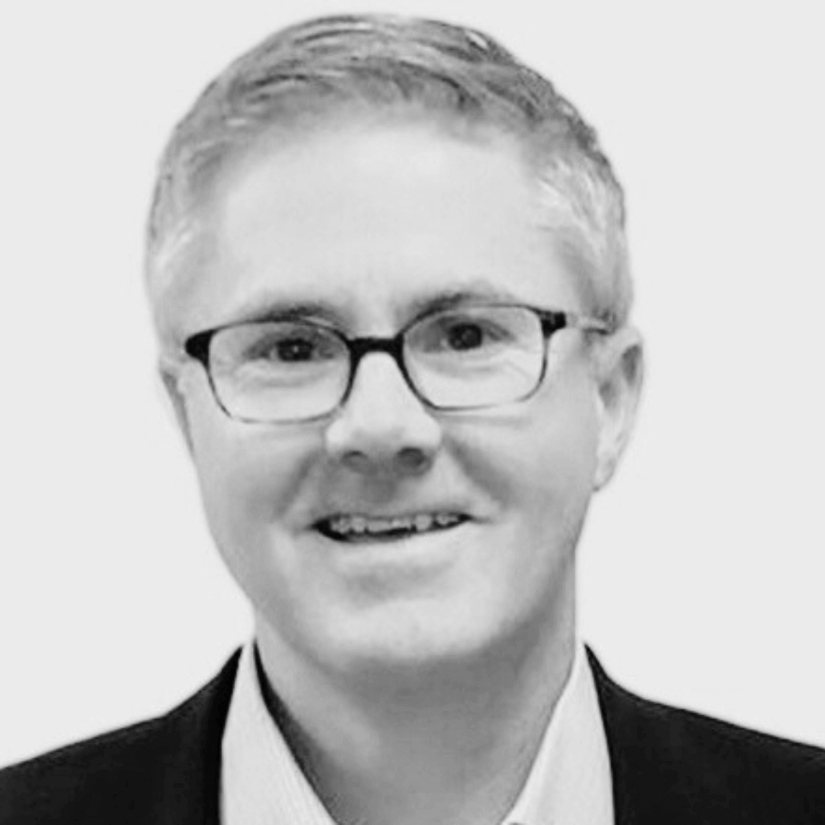 Black and white headshot of a smiling man with short hair and glasses. He is wearing a suit jacket and collared shirt against a plain background.